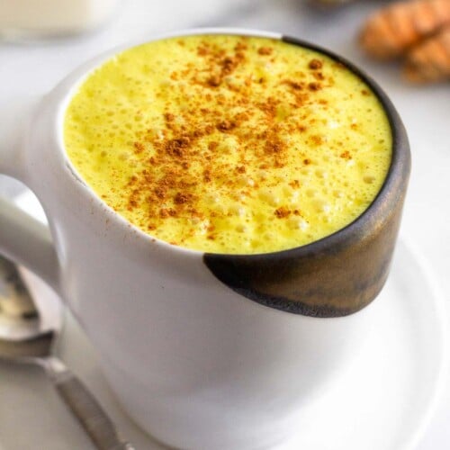 Golden milk latte sprinkled with cinnamon in a white mug sitting on a plate with a spoon on it. Behind it are pieces of fresh turmeric and ginger and a glass jar of milk.