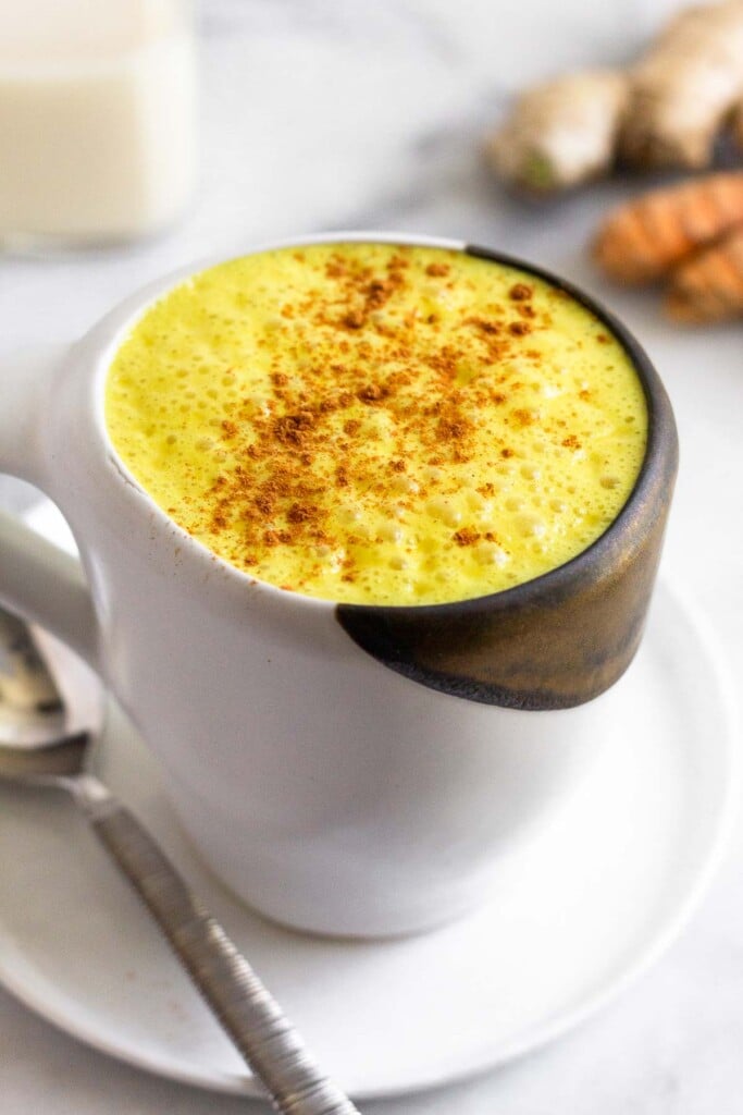 Golden milk latte sprinkled with cinnamon in a white mug sitting on a plate with a spoon on it. Behind it are pieces of fresh turmeric and ginger and a glass jar of milk.