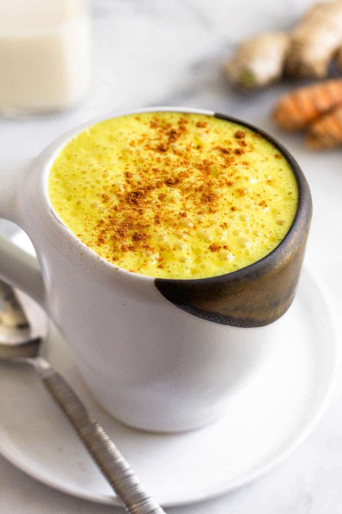 Golden milk latte sprinkled with cinnamon in a white mug sitting on a plate with a spoon on it. Behind it are pieces of fresh turmeric and ginger and a glass jar of milk.