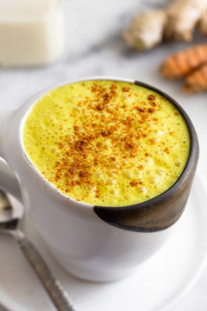 Turmeric milk sprinkled with cinnamon in a white mug on a plate with a spoon on it.