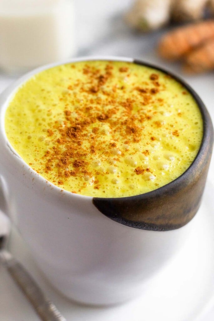 Close up of a mug filled with a golden milk latte sprinkled with cinnamon. Next to the mug is piece of silverware and behind it is a piece of fresh turmeric, a piece of fresh ginger, and a jar of milk.