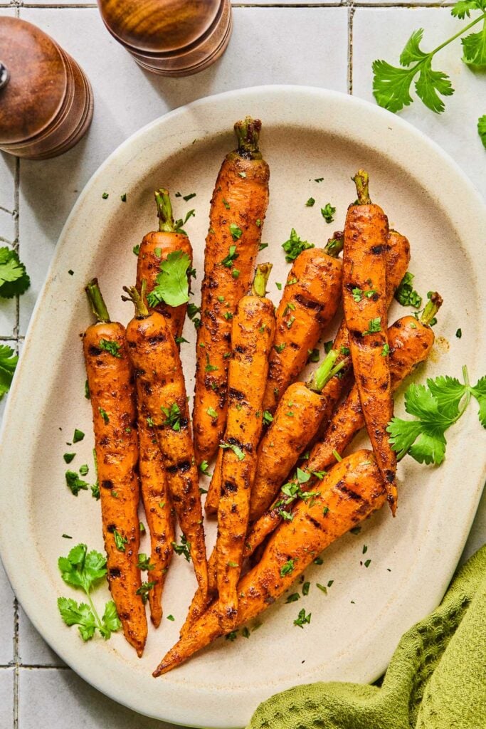 A pile of grilled carrots topped with fresh cilantro on a serving platter. Around the platter is a green kitchen towel, more fresh cilantro, and a salt and pepper shaker.