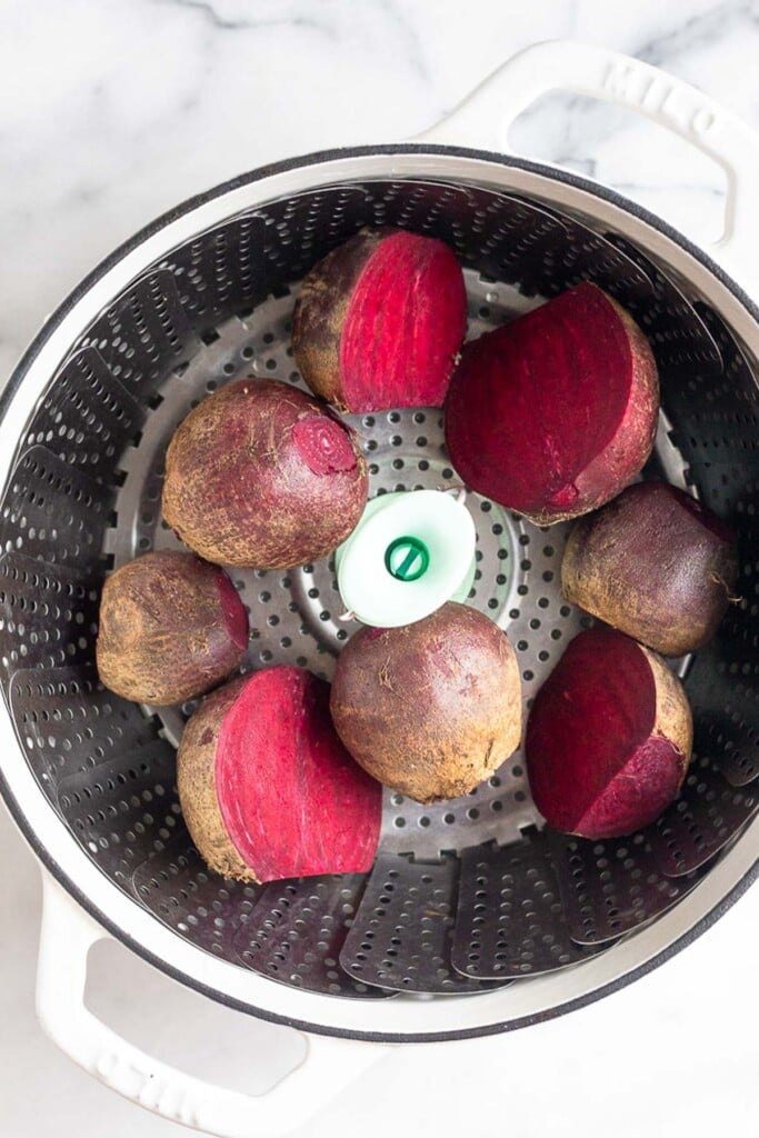 Raw red beets in a large pot with a steamer basket in it. Some of the beets are sliced in half.