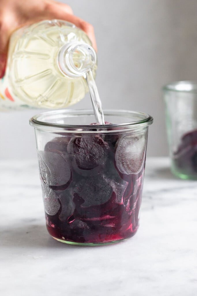 Jar filled with sliced cooked beets with white vinegar being poured in it. Behind the jar is another jar filled with more sliced beets.