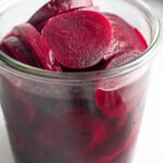 A jar of pickled beets. Behind the jar in the background is a raw red beet and in front of the jar is a striped kitchen towel.
