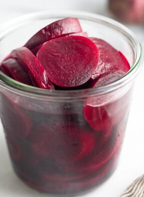 A jar of pickled beets. Behind the jar in the background is a raw red beet and in front of the jar is a striped kitchen towel.