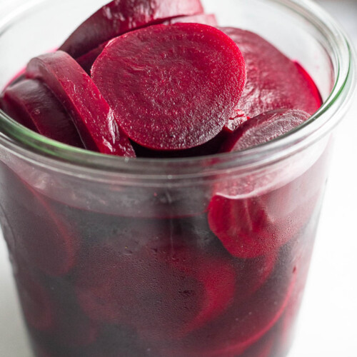 A jar of pickled beets. Behind the jar in the background is a raw red beet and in front of the jar is a striped kitchen towel.