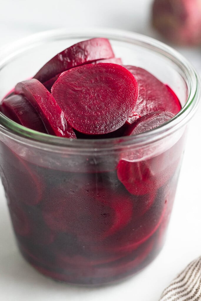 A jar of pickled beets. Behind the jar in the background is a raw red beet and in front of the jar is a striped kitchen towel.