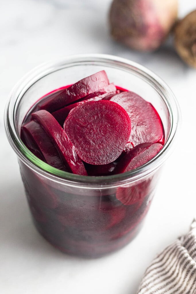 A large glass jar of refrigerator pickled beets. Behind the jar is two raw red beets and in front of the jar is a striped kitchen towel.