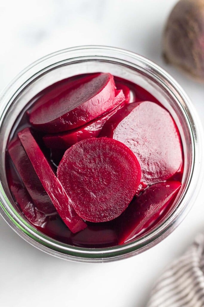 Overhead shot of a jar of quick pickled beets. Next to the jar is a raw red beet and a striped kitchen towel.