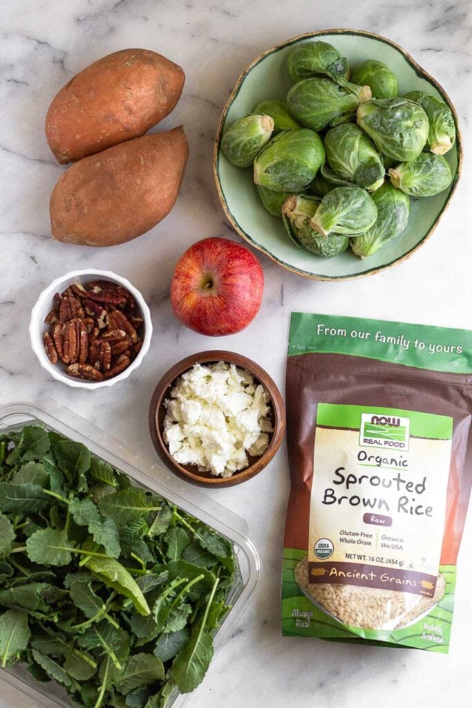 White marble counter with a bowl of raw brussel sprouts, a red apple, a bag of brown sprouted rice, a bowl of goat cheese, a plastic container of baby kale, a bowl of pecans, and two sweet potatoes.