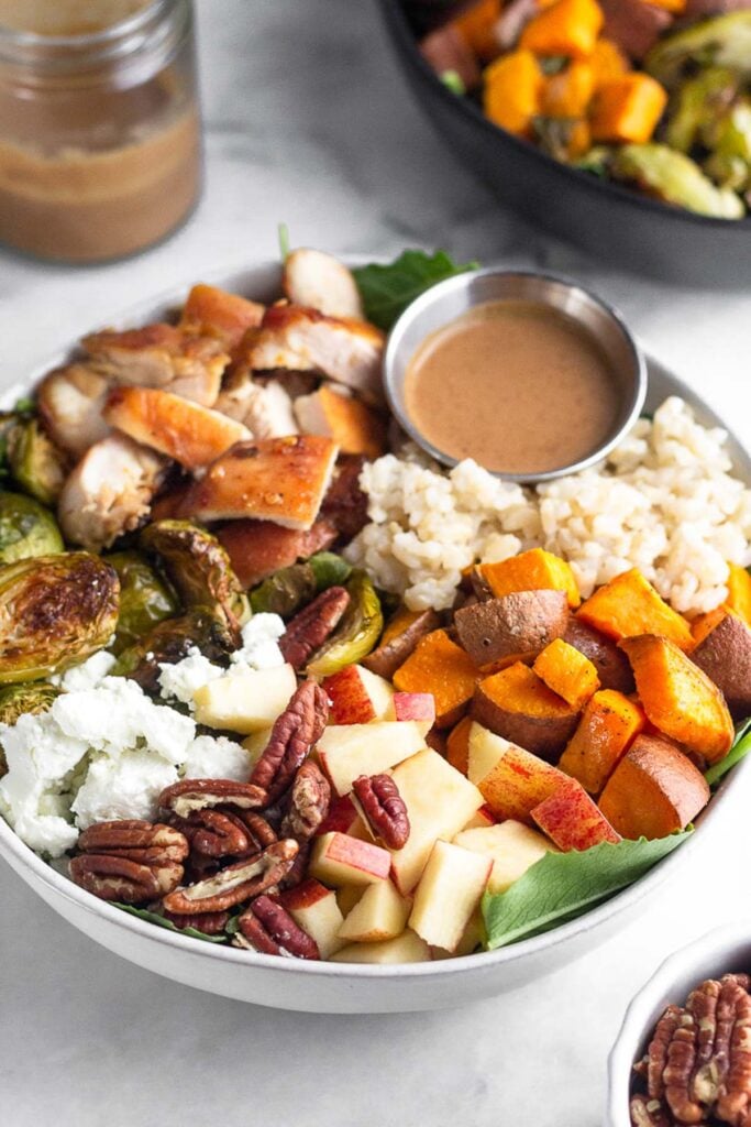 Sweetgreen harvest salad in a white bowl with rice, roasted sweet potatoes, diced apples, pecans, goat cheese, roasted brussel sprouts, diced chicken, and a ramekin of balsamic dressing over greens. In front of the bowl is a small bowl of pecans and behind the bowl is another bowl of salad and jar of dressing.