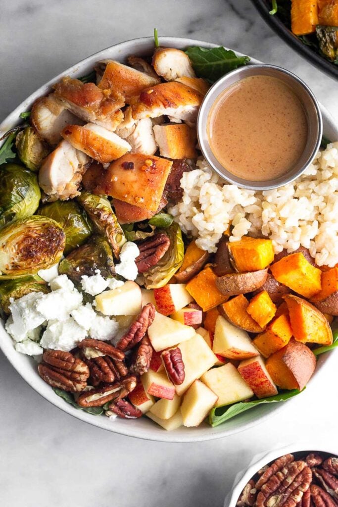 Copycat sweetgreen harvest bowl with rice, roasted sweet potatoes, diced apples, pecans, goat cheese, roasted brussel sprouts, diced chicken, and a small ramekin of creamy balsamic dressing over greens. Next to it is a bowl of pecans.