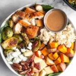 Sweetgreen harvest bowl with rice, roasted sweet potatoes, diced apples, pecans, goat cheese, roasted brussel sprouts, diced chicken, and small ramekin of creamy balsamic dressing over greens. Another harvest bowl is off to the side and a small bowl of pecans to the other side.