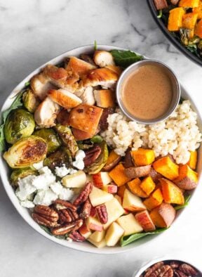 Sweetgreen harvest bowl with rice, roasted sweet potatoes, diced apples, pecans, goat cheese, roasted brussel sprouts, diced chicken, and small ramekin of creamy balsamic dressing over greens. Another harvest bowl is off to the side and a small bowl of pecans to the other side.