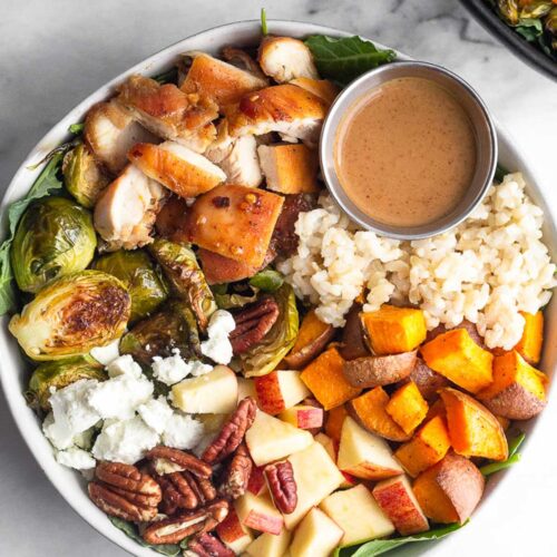 Sweetgreen harvest bowl with rice, roasted sweet potatoes, diced apples, pecans, goat cheese, roasted brussel sprouts, diced chicken, and small ramekin of creamy balsamic dressing over greens. Another harvest bowl is off to the side and a small bowl of pecans to the other side.