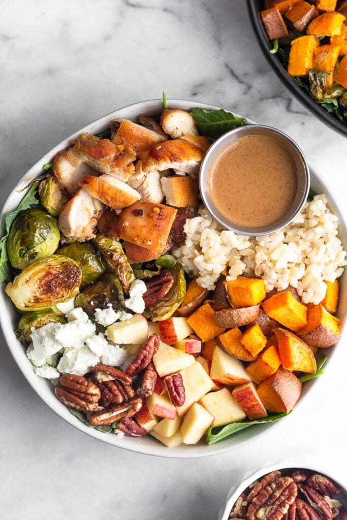 Sweetgreen harvest bowl with rice, roasted sweet potatoes, diced apples, pecans, goat cheese, roasted brussel sprouts, diced chicken, and small ramekin of creamy balsamic dressing over greens. Another harvest bowl is off to the side and a small bowl of pecans to the other side.