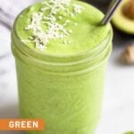 Green protein smoothie in a jar topped with coconut flakes and a metal straw in the jar. Around it is half a lime, a piece of ginger root, half an avocado, and a bowl of frozen pineapple.