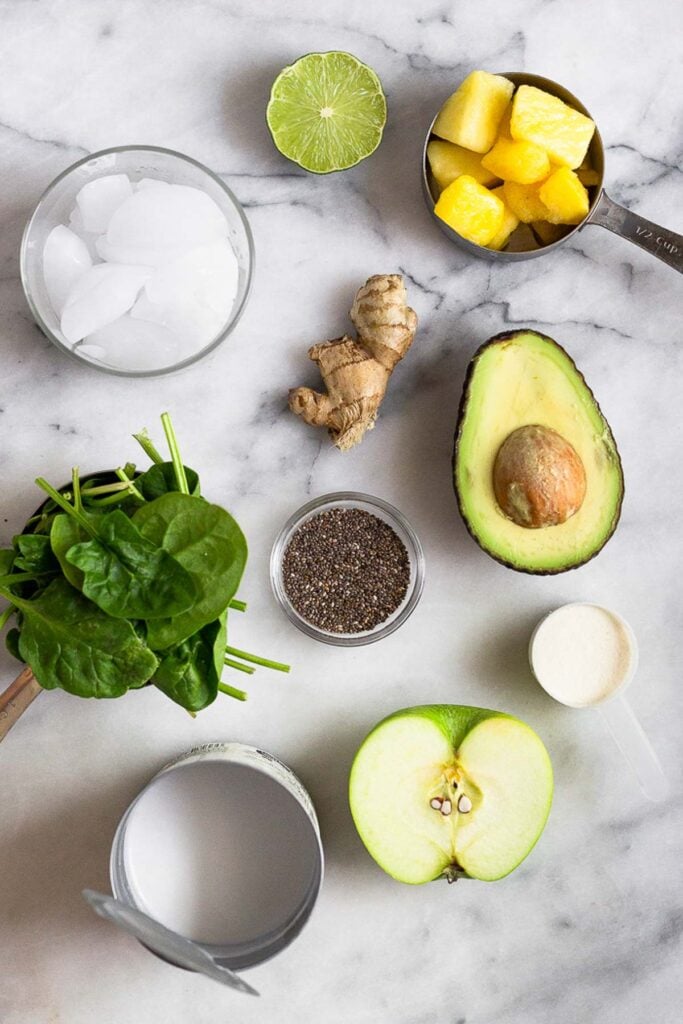 Overhead shot of a white marble counter with a bowl of frozen pineapple, some ginger root, a bowl of chia seeds, half an avocado, a scoop of protein powder, half a green apple, a can of coconut milk, a cup of spinach, a glass of ice, and half a lime.