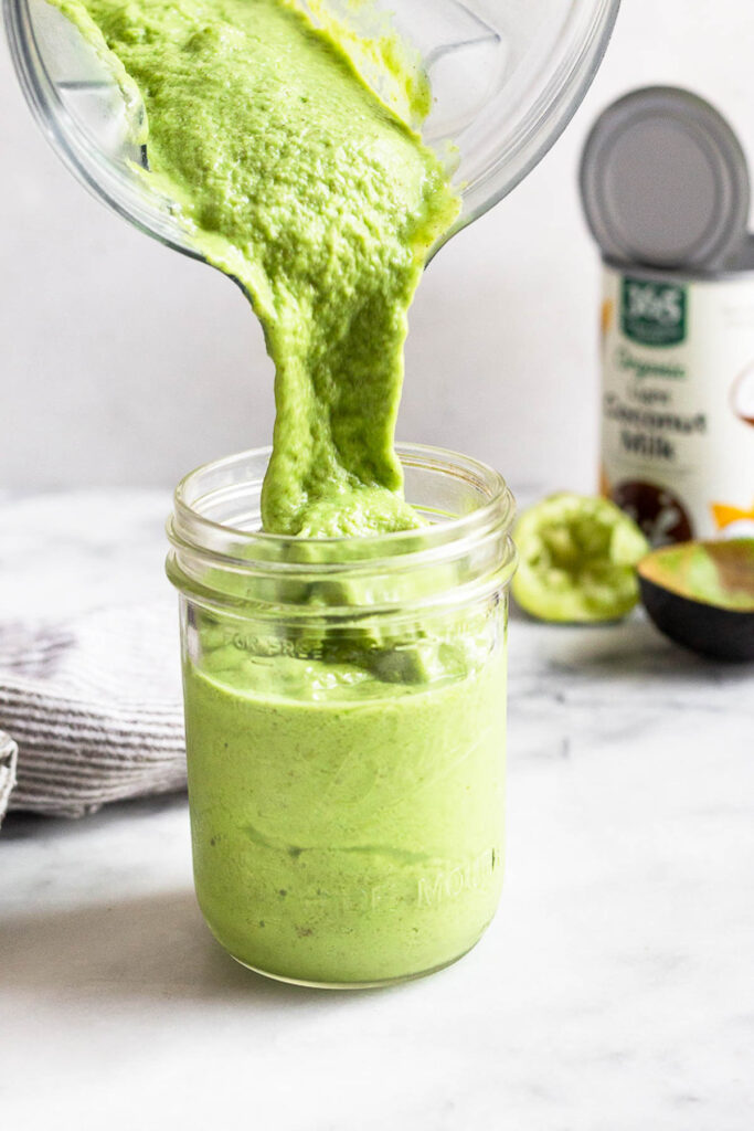 Protein green smoothie being poured into a glass jar from a blender. Behind it is a can of coconut milk, half a lime, and an empty half of avocado.