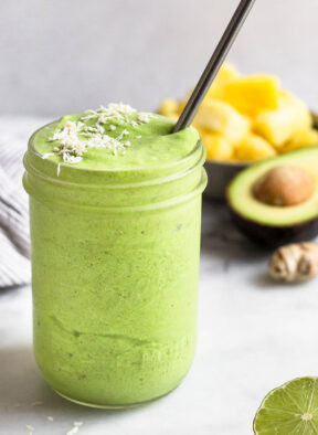 Green protein smoothie in a jar topped with coconut flakes and a metal straw in it. Around it is half a lime, half an avocado, ginger root, and a bowl of frozen pineapple.
