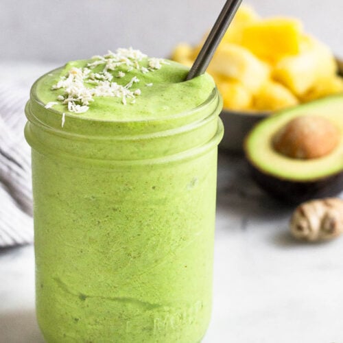 Green protein smoothie in a jar topped with coconut flakes and a metal straw in it. Around it is half a lime, half an avocado, ginger root, and a bowl of frozen pineapple.