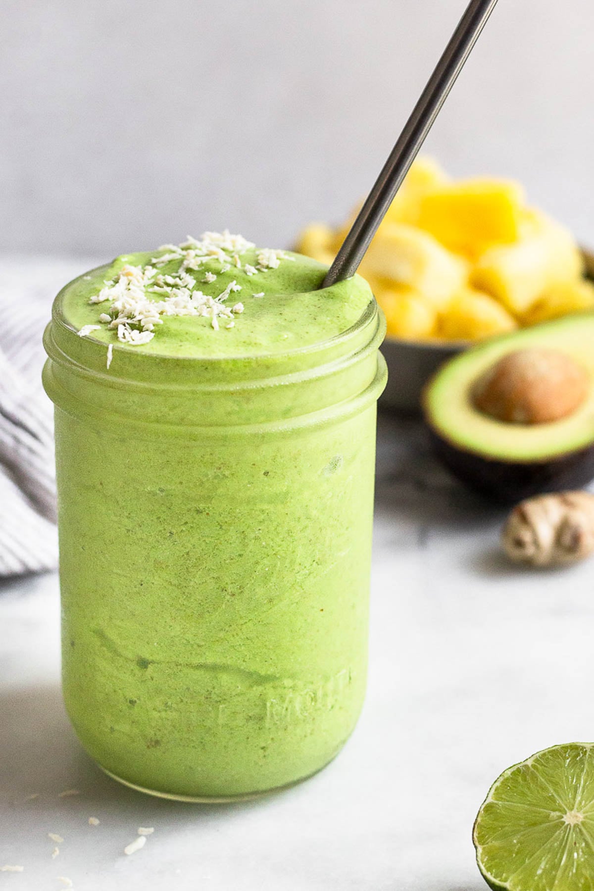 Green protein smoothie in a jar topped with coconut flakes and a metal straw in it. Around it is half a lime, half an avocado, ginger root, and a bowl of frozen pineapple.