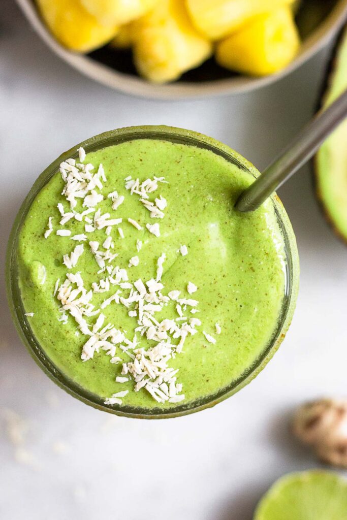 Overhead shot of a green smoothie topped with shredded coconut and a straw in it. Around it a bowl of frozen pineapple, half an avocado, ginger root, and half a lime.