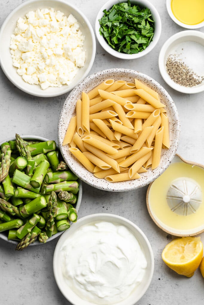 Grey concrete counter with a bowl of olive oil, a bowl of chopped herbs, a bowl of salt and pepper, and bowl of uncooked penne, a ceramic juicer with lemon juice in it, a juiced lemon, a bowl of greek yogurt, a bowl of chopped asparagus, and a bowl of feta.