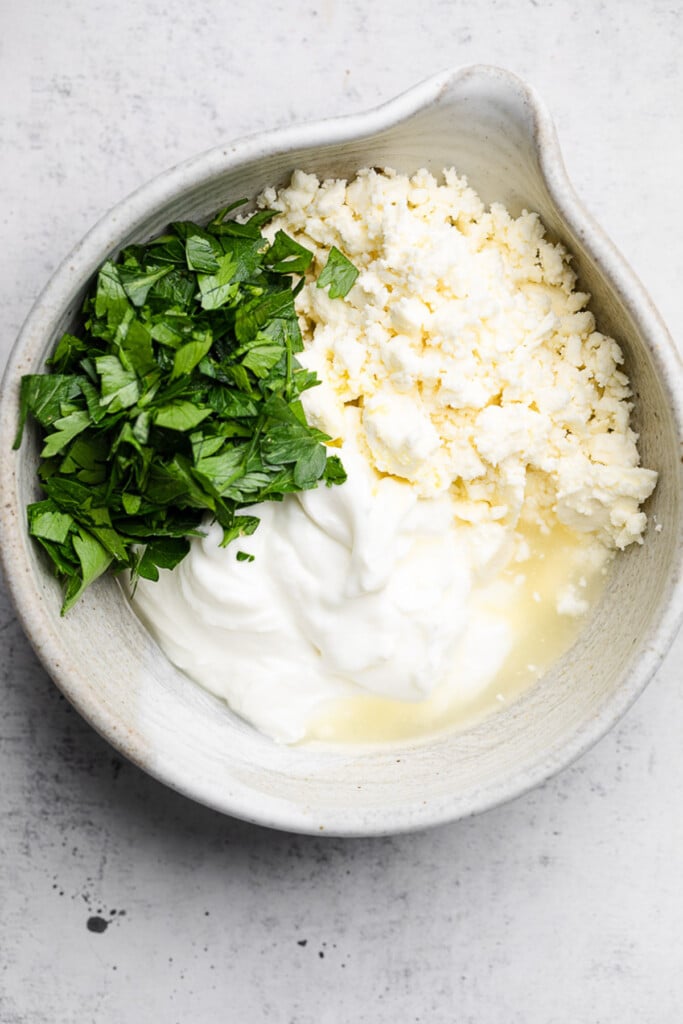 A white mixing bowl with greek yogurt, feta, chopped herbs, and olive oil in it before it is mixed together.