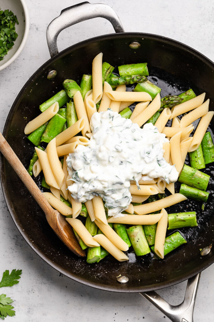 Skillet filled with chopped sautéed asparagus, cooked penne, and a thick and creamy white sauce before it is mixed together. A wooden spoon is in the pan as well.