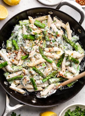 Large skillet filled with asparagus pasta in a creamy feta sauce. It is topped with lemon zest and red peppers flakes and a wooden spoon is in the bowl. Around the pan is a dish of chopped herbs, some lemons, a bowl of feta, and a small dish of red pepper flakes.