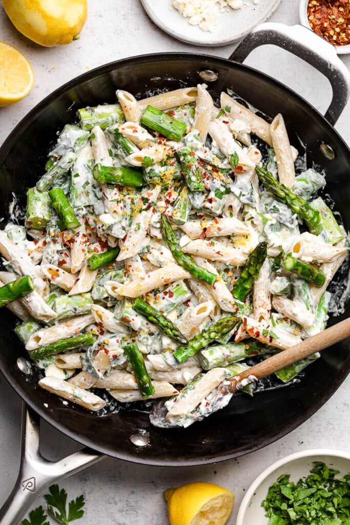 Large skillet filled with asparagus pasta in a creamy feta sauce. It is topped with lemon zest and red peppers flakes and a wooden spoon is in the bowl. Around the pan is a dish of chopped herbs, some lemons, a bowl of feta, and a small dish of red pepper flakes.