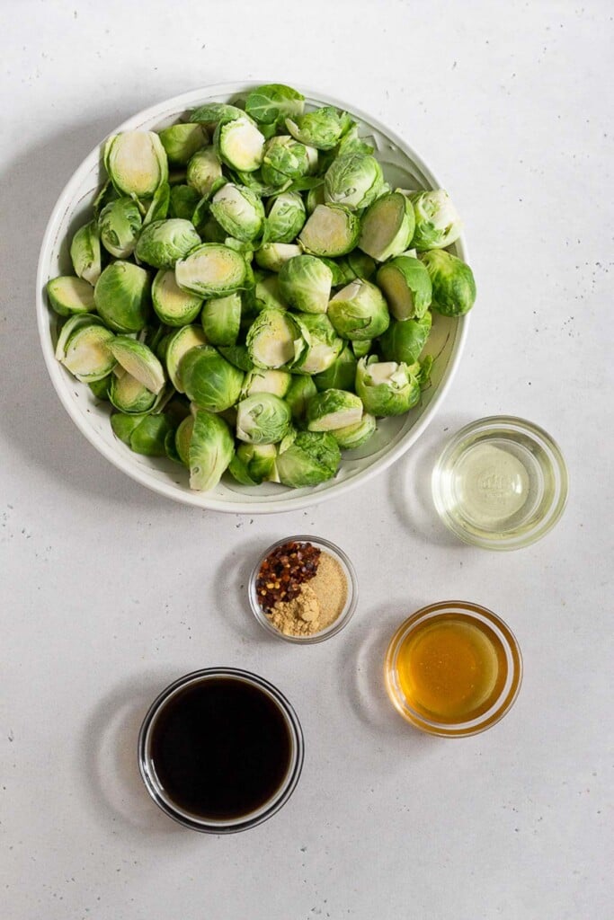 White counter with a bowl of raw halved brussel sprouts, a small bowl of oil, a bowl of honey, a bowl of coconut aminos, and a bowl of spices.