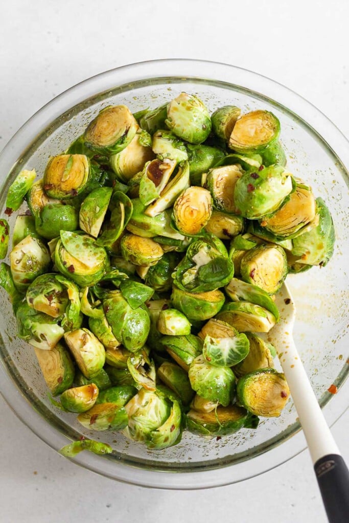 Large glass bowl filled with raw halved brussel sprouts coated in oil and spices. There is a spatula in the bowl as well.