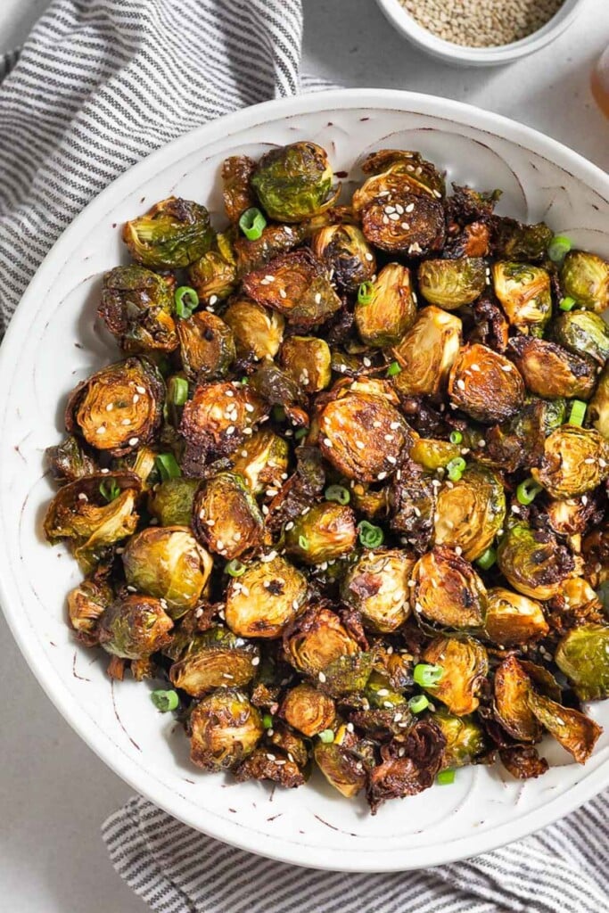Crispy air fryer brussel sprouts topped with sesame seeds and sliced greens onions in a large white bowl. The bowl is sitting on a striped towel with a bowl of sesame seeds next to it.