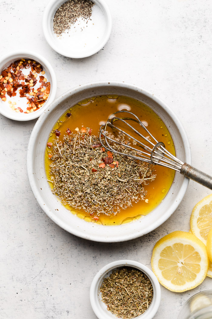 A small bowl with pasta salad vinaigrette in it with a small whisk. Next to it is small bowls of spices and some sliced lemons.