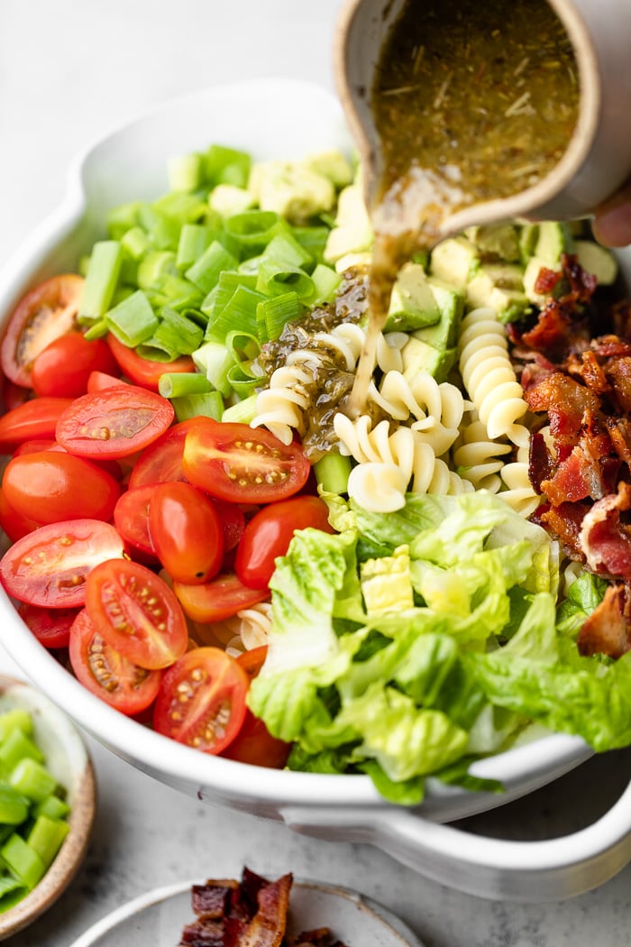 Bacon pasta salad in a large bowl with homemade pasta salad dressing being poured over top.