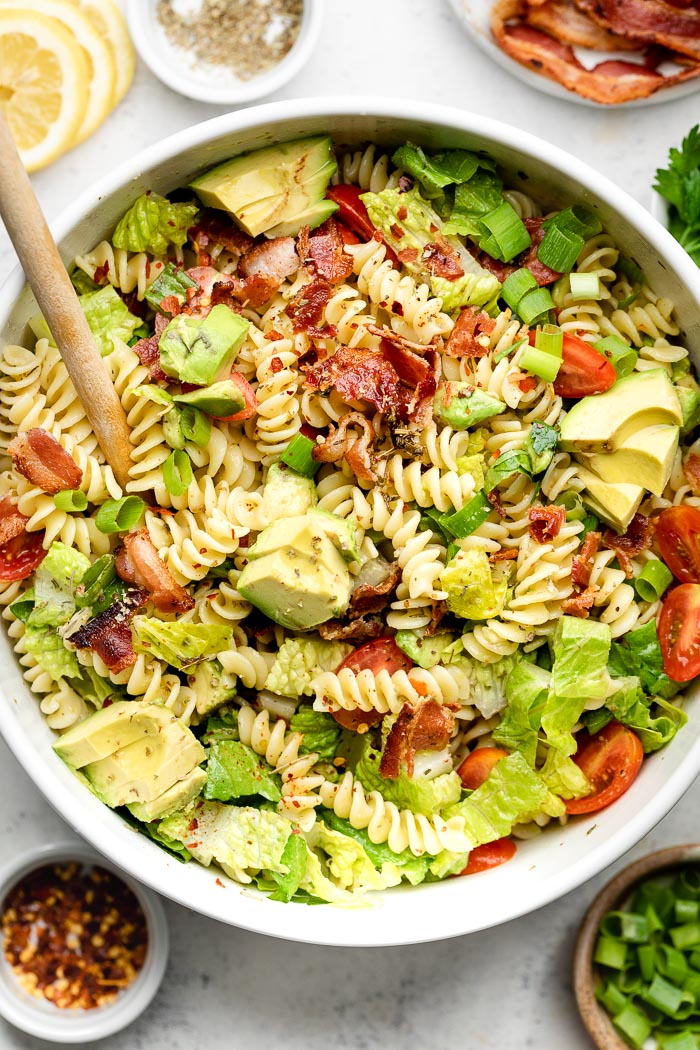 BLT pasta salad with Italian dressing and bacon in a large white bowl with a wooden spoon coming out of it. Around it is a few dishes of herbs, lemon, spices, and green onions.