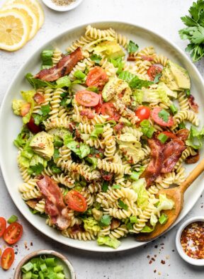 Blt pasta salad on a large white dish and a spoon in it. Around it is s