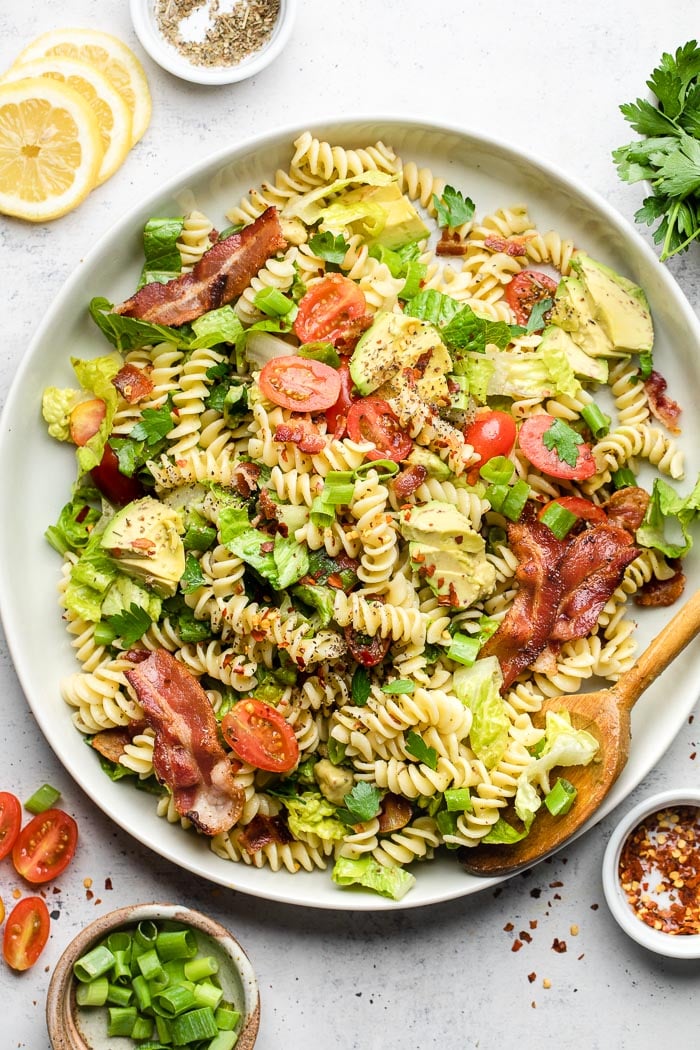 Blt pasta salad on a large white dish and a spoon in it. Around it is s
