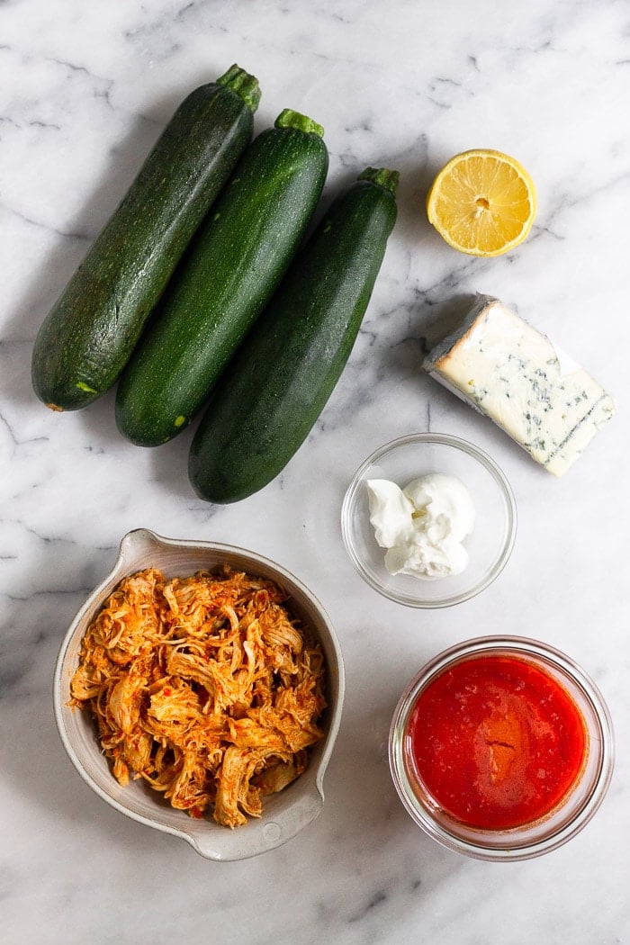 White counter top with 3 zucchinis, half a lemon, block of blue cheese, bowl of yogurt, jar of buffalo sauce, and a bowl of shredded buffalo chicken.