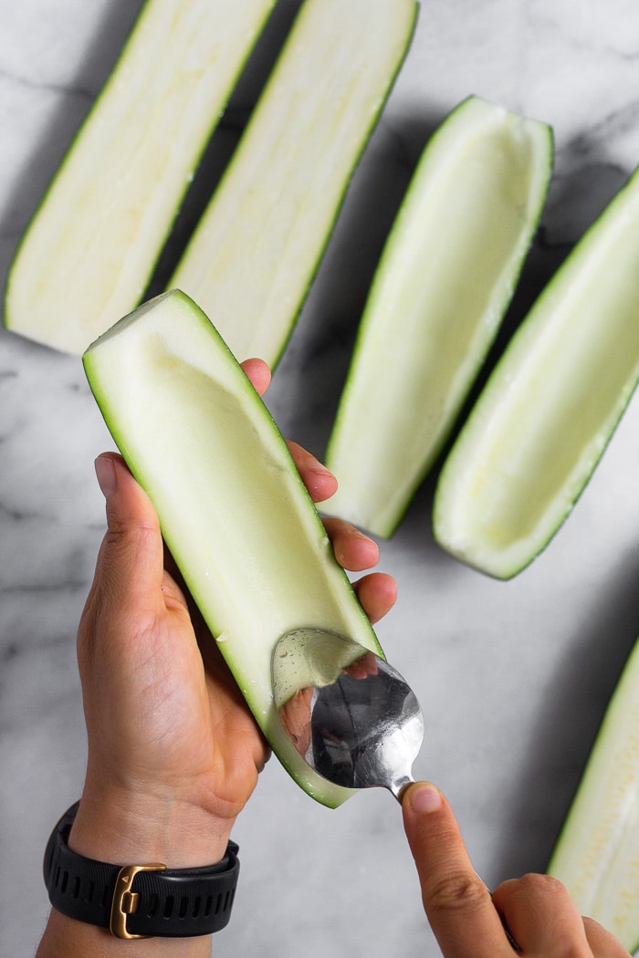 Overhead shot of a hand holding half a zucchini that is cut lengthwise. It is scooping out the inside flesh with a spoon. There are more zucchini in the background.
