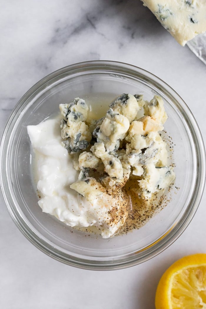 Overhead shot of a small glass bowl with yogurt, blue cheese, lemon juice, and salt and pepper. Next to it is more blue cheese and half a lemon wedge.