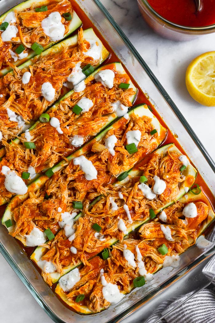 Overhead shot of buffalo chicken stuffed zucchini boats topped in a baking dish. They are topped with blue cheese dressing and green onions. Next to the pan is half a lemon wedge and a jar of buffalo sauce.