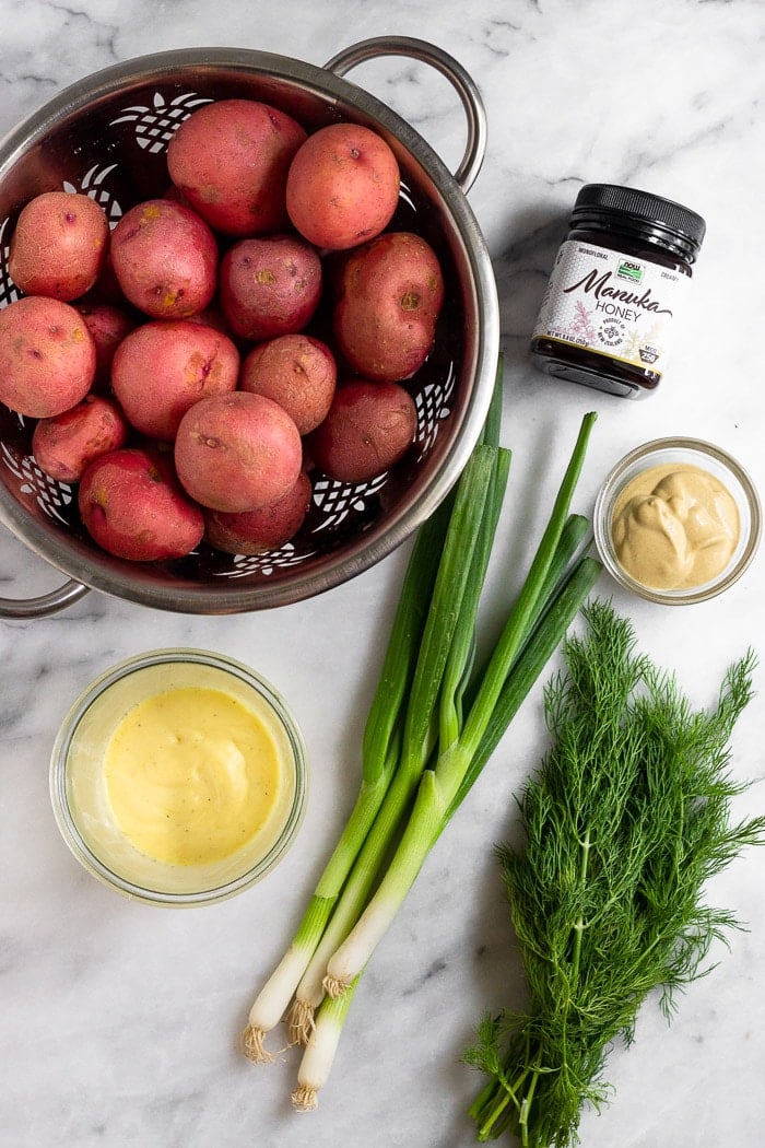 White counter top with a colander of red potatoes, jar of honey, bowl of mustard, fresh dill, green onions, and a jar of mayo.