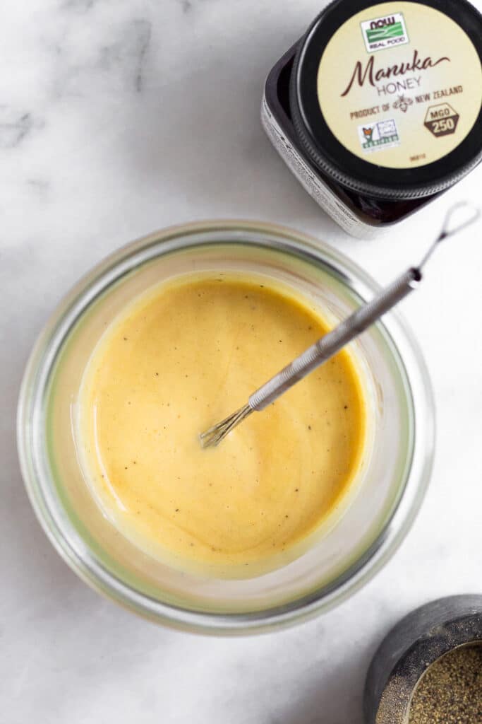 Healthy honey mustard dressing in a jar with a small whisk in it. Next to is a jar of honey and a small dish of pepper.
