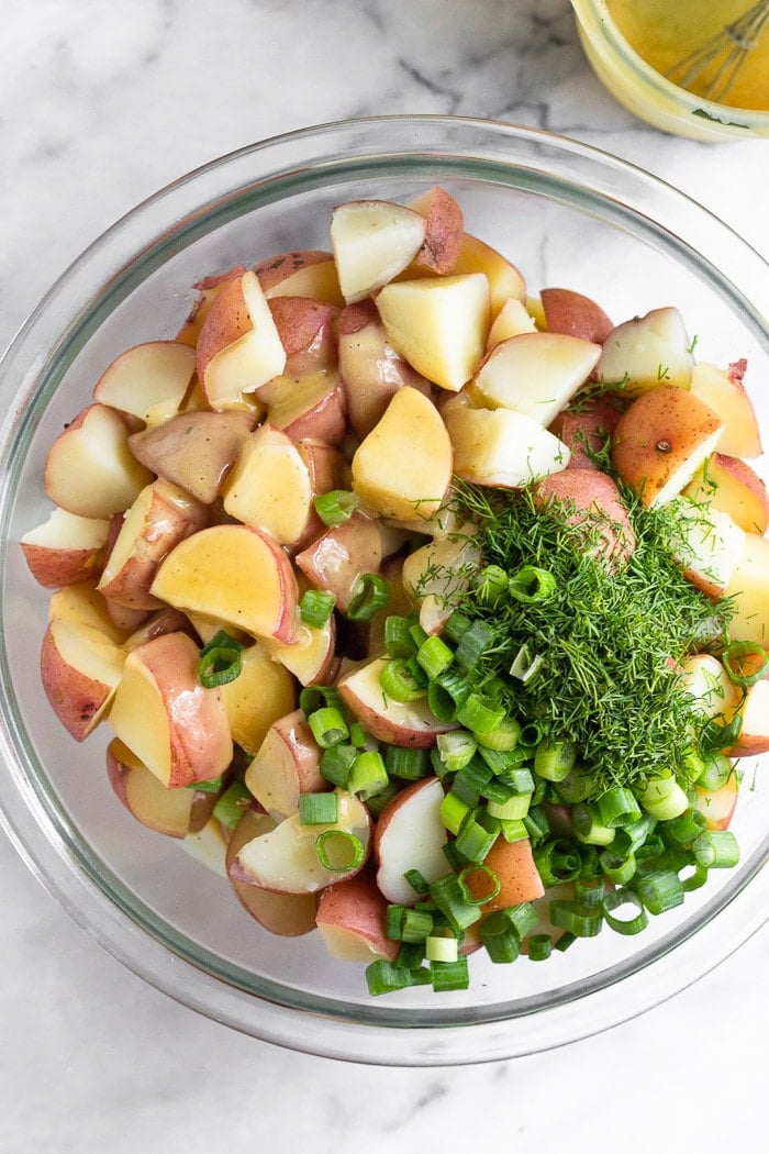 Overhead shot of a large glass bowl filled with cooked cubed red potatoes with honey mustard dressing, dill, and green onions over top.