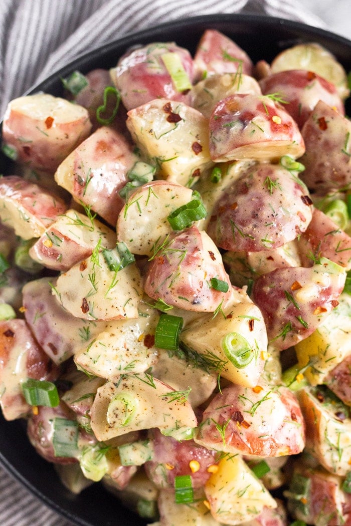 Overhead shot of healthy red potato salad with topped with green onions and red pepper flakes. It is in a black bowl with a striped kitchen towel underneath.