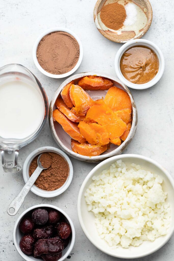 White counter with a plate of spices, a bowl of almond butter, a bowl of frozen sweet potatoes, a bowl of frozen cauliflower rice, a bowl of frozen cherries, a dish of cocoa powder, a measuring cup of milk, and a bowl of chocolate protein powder.